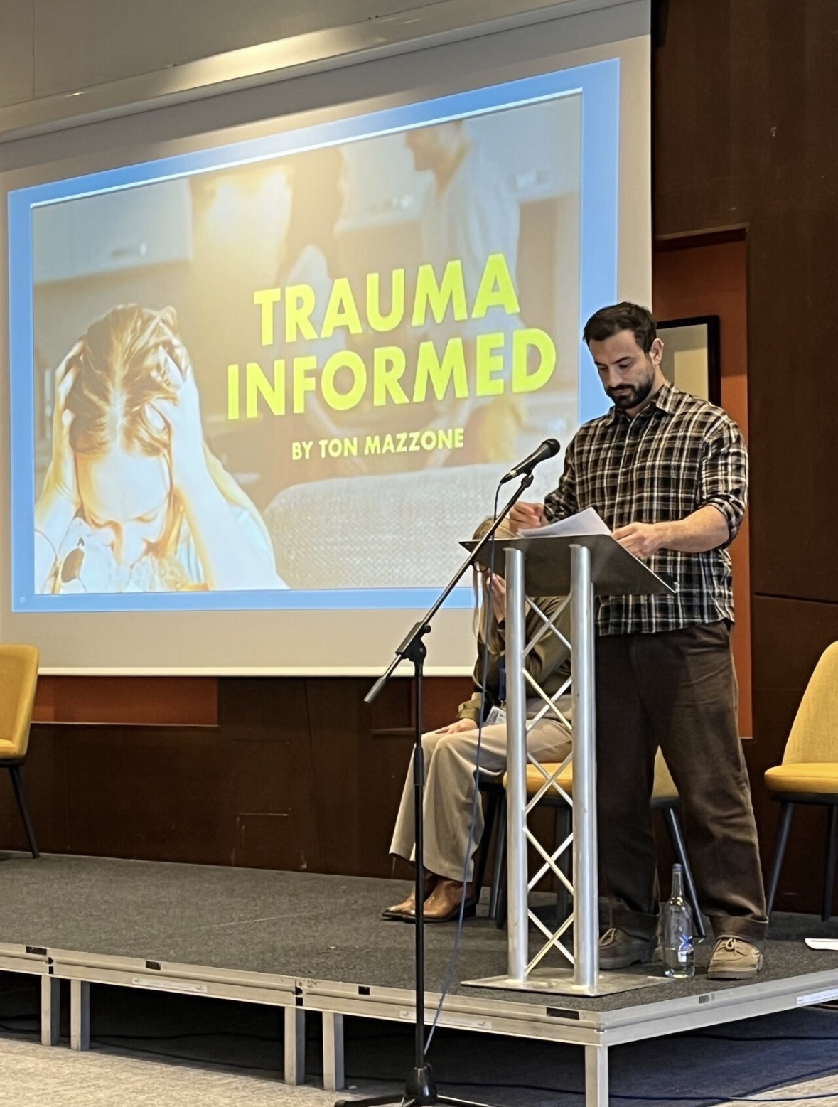 Ton Mazzone reading his poem Trauma Informed at the lectern, with the title on the screen behind him